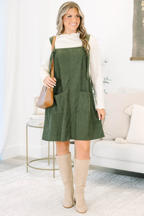 Plus size olive green corduroy overall dress with tie straps and front pockets over a cream top.