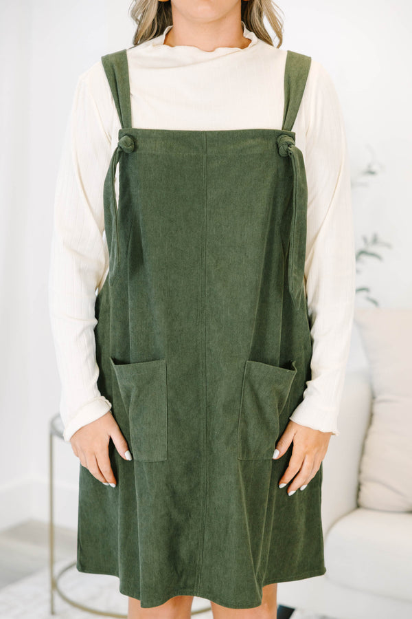 Plus size olive green corduroy overall dress with tie straps and front pockets over a cream top.