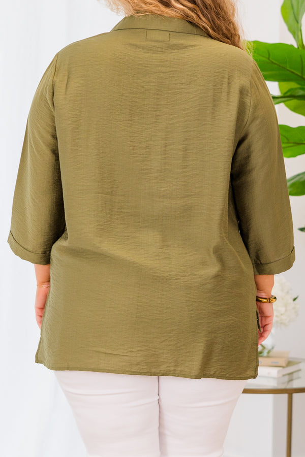 A woman smiles wearing a button-down olive green top with three-quarter sleeves and white pants.