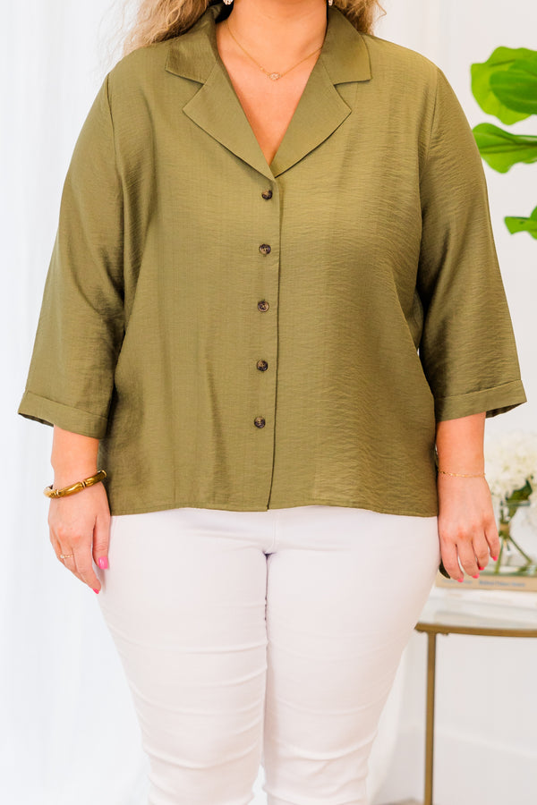 A woman smiles wearing a button-down olive green top with three-quarter sleeves and white pants.