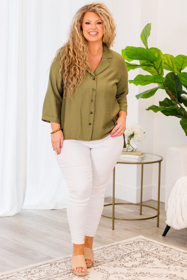 A woman smiles wearing a button-down olive green top with three-quarter sleeves and white pants.