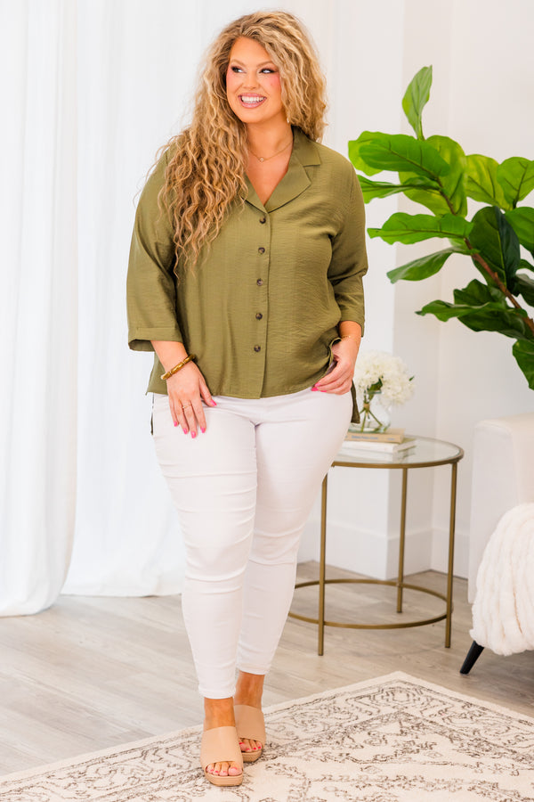A woman smiles wearing a button-down olive green top with three-quarter sleeves and white pants.
