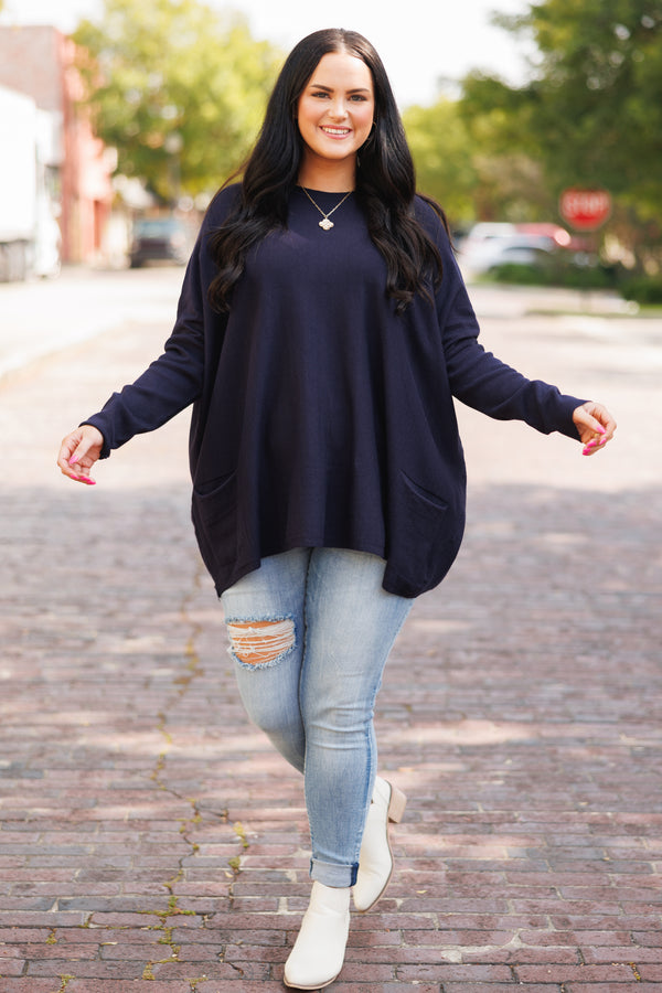 A woman smiles wearing a long-sleeved navy blue tunic with front pockets and light-wash jeans.